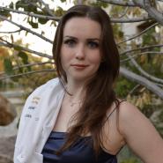 Jillian Cellucci with University of California Davis lab coat over her shoulder, wearing a blue dress and standing by a tree.