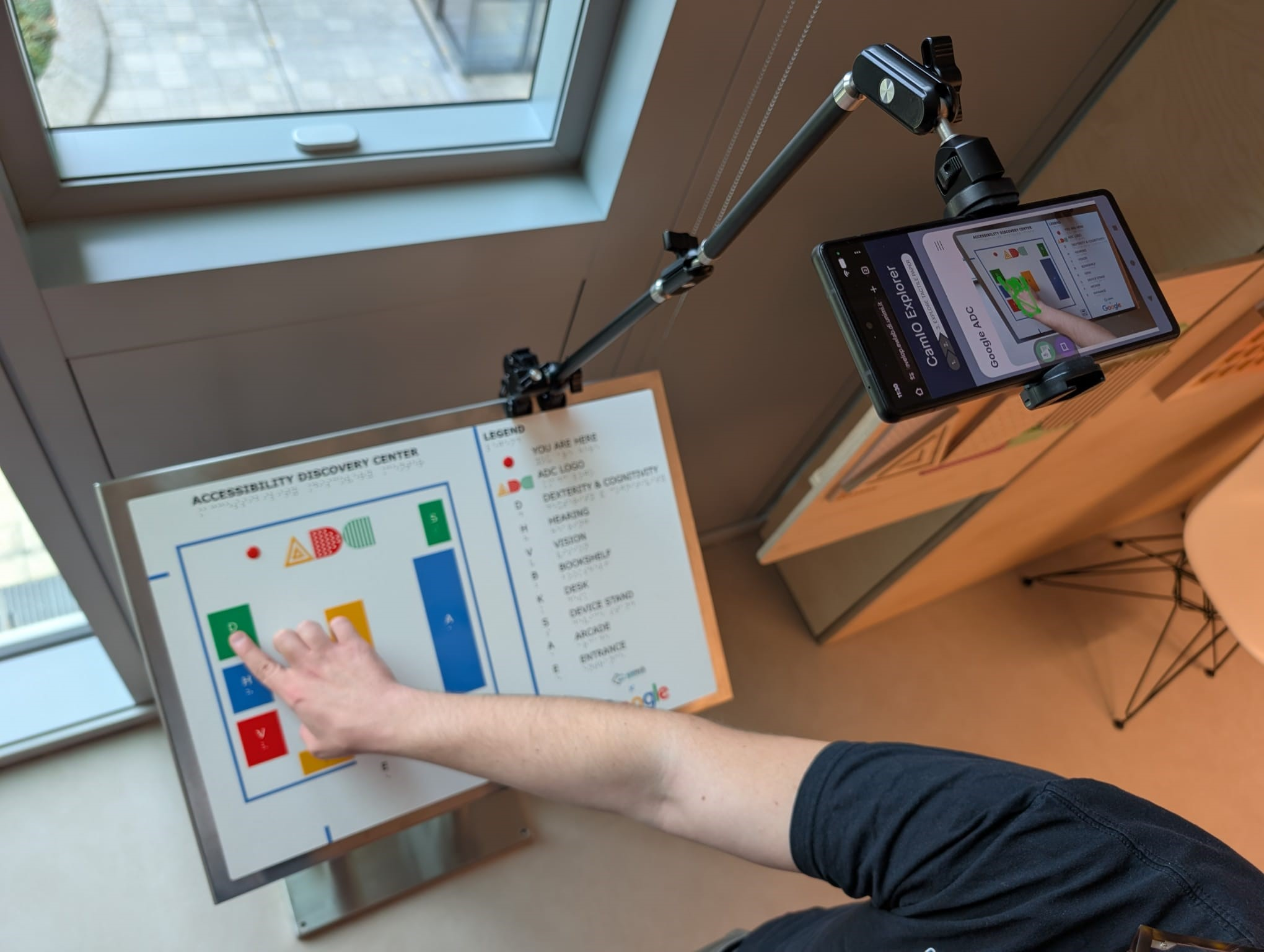 User pointing with index finger to a feature on tactile map, with Android smartphone mounted above with screen showing view of tactile map and user's arm.